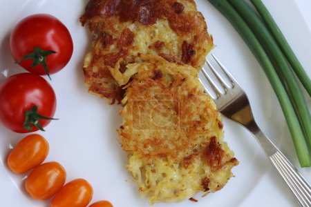 Closeup of delicious homemade fried grated potato pancakes served with fresh tomatoes and green onions on the white plate, fork on the right side. Draniki. Vegetable meal. Top view.
