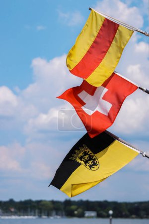 Téléchargez les photos : Trois drapeaux sur les mâts brandissant dans le vent. Drapeaux de Bade, Suisse, Drapeau Bade-Wrttemberg avec un emblème. Drapeau suisse au milieu. Isolé sur le ciel bleu et les nuages blancs. Espace de copie. - en image libre de droit