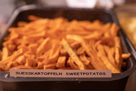 Photo for Yummy French fries in the metal tray with the tag Suesskartoffeln Sweetpotatos. Potato chips on the counter at the street market. Product Sign at market. Selective focus. Side view. Bokeh. - Royalty Free Image