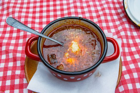 Téléchargez les photos : Soupe de chou russe ukrainienne slovaque traditionnelle avec saucisse et viande fumée dans un pot en métal rouge rétro servi sur l'assiette avec une cuillère. Bol de délicieux Soljanka borch à la crème sure blanche. Vue du dessus - en image libre de droit