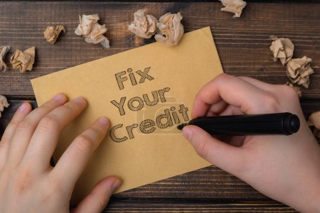 A person is writing on a piece of paper that says Fix Your Credit. The paper is on a wooden surface and there are some shredded pieces of paper around it