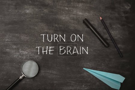 Turn on the brain is written on the chalkboard. The chalkboard is covered with chalk and a pair of pencils. A blue paper airplane is also on the chalkboard