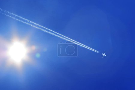 a passenger plane with contrails in the blue sky with sun and sunbeams