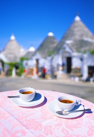 two cups of Espresso on a table with pink tablecloth, with empty street and souvenir stores out of focus in background in Alberobello in preseason in April, with Trulli, blue sky, Alberobello, Apulia, Italy