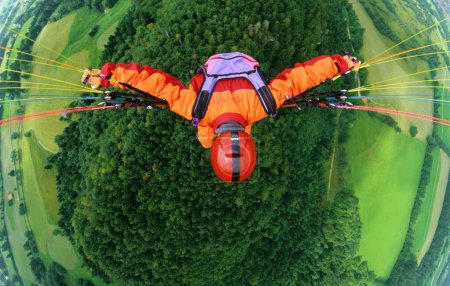 Paraglider pilot with orange clothes and red helmet flying in 500 ft above a forest and meadows, bird's eye view, Brauneck, Lenggries, Bavaria, Germany