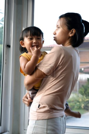 Asiatin hält ihr Baby im Arm, während sie zu Hause neben einem Fenster steht.