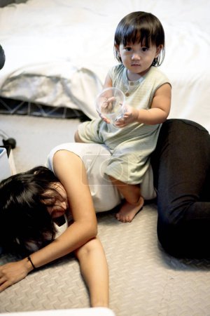 Exhausted mother lies on floor as son plays on top of her. Exhausted mothers experience postpartum depression.