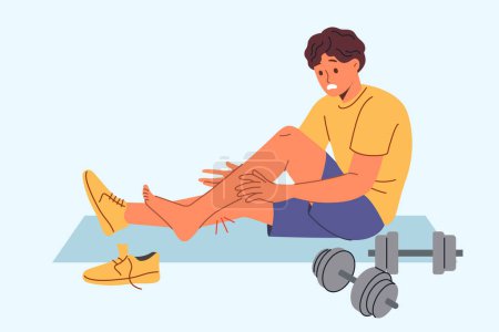Injured sportsman sits on mat for sports and gymnastics near dumbbells, after stretching muscles on legs. Injured athlete experiences pain and agony after careless use of sports equipment