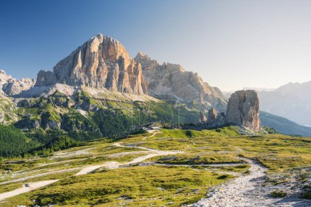 Cinque Torri peaks and Tofane Mountains in Italian Dolomites at summer cloudless morning with green grass lighteen by warm light of sun from right side and path inf front
