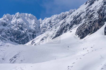 Trekking y skituring en las altas montañas de Tatra en invierno. Sendero en la nieve con la gente y las montañas rocosas gigantes en el bacground en día soleado con cielo azul claro.