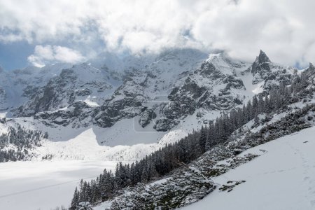 Winter Tatra mountains panorama with snowy trees and frozen Morkie Oko. High rocky mountains covered with clouds lighteen by sun and Mnich (Monk) peak in the background.