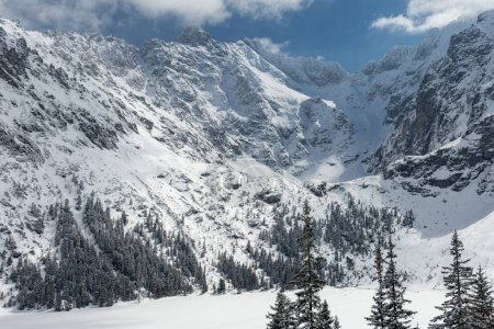 Montañas polacas de Tatra en invierno con árboles nevados y altos picos rocosos congelados en el fondo con cielo azul y algunas nubes en el día soleado. Sendero visible a la vaina Czarny Staw Rysami.