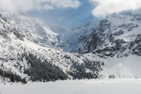 Frozen Morskie Oko Lake covered with snow in the sunny day with people on its surface. Partly cloud covered High Tatra Mountains peaks like Rysy in the background.
