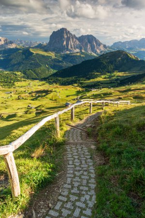View from Seceda mountain towards the Sassolungo mouuntain in the Italian Dolomites in sunny sumer day with path and wooden fence in foreground nad green grass and blue sky with clouds.