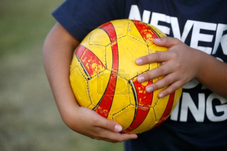 Child playing soccer. Boy holding a soccer ball in his arms. Close up. Play soccer. Sport. Childhood. Old yellow and red ball very used. Fun, summer, vacation, outdoor entertainment.
