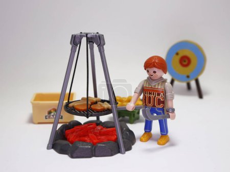 Photo for Playmobil. Man doing a barbecue in a camp. Boy grilling hot dogs. Bonfire in camp. Preparing a picnic lunch. Redhead. Sausages. Toy for children. Doll and accessories. - Royalty Free Image
