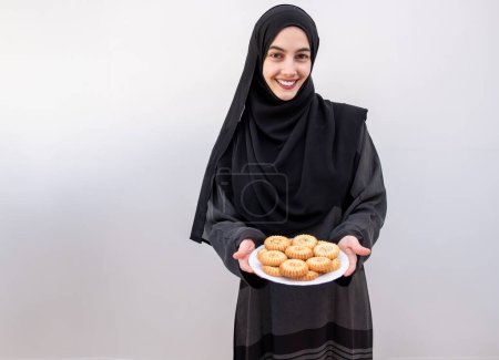 Radiante alegría y orgullo cultural, una mujer árabe en abaya delicadamente sostiene los preciados dulces del Eid, Maamoul, sobre un sereno fondo blanco, personificando el espíritu de la celebración del Ramadán y el Eid.