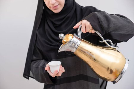 Exudando calidez y tradición, una mujer árabe presenta elegantemente la preciada cafetera y taza árabe, un símbolo de hospitalidad y alegría durante el Ramadán y el Eid, sobre un sereno telón de fondo blanco