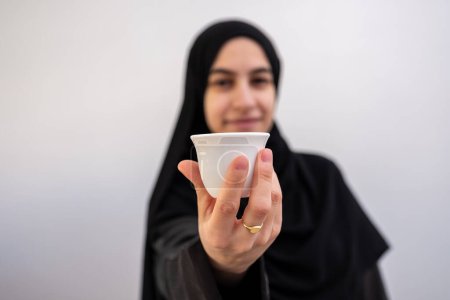 Exudando calidez y tradición, una mujer árabe presenta elegantemente la preciada cafetera y taza árabe, un símbolo de hospitalidad y alegría durante el Ramadán y el Eid, sobre un sereno telón de fondo blanco