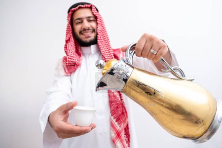 Hombre árabe sosteniendo cafetera árabe usando dishdasha y kandura sobre fondo blanco aislado con generosidad y sentimientos de hospitalidad