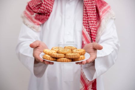 In a gesture of hospitality and generosity, an Arabic man proudly presents a plate of Maamoul, symbolizing the warmth and abundance of Eid celebrations. Clad in the traditional Kandura and Keffiyeh