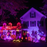 Night photo. Glowing Halloween decorations - skeletons, witches, pumpkins around a small wooden house.