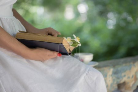Téléchargez les photos : Mains de femme avec un livre et une fleur de jasmin dans un jardin verdoyant. Une tasse de thé en arrière-plan sur le parapet du gazebo. - en image libre de droit
