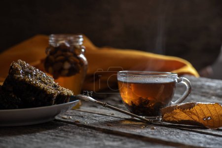 Foto de Foto oscura en un estilo rústico. Taza de té con humo, miel y panal sobre un fondo de madera áspera. El estado de ánimo de un cálido otoño hogareño. - Imagen libre de derechos