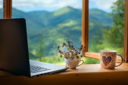 Laptop und eine Tasse Tee auf der Fensterbank eines offenen Fensters mit Blick auf eine herrliche Berglandschaft