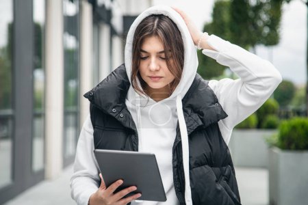 Foto de Mujer joven de negocios se para fuera y utiliza una tableta contra un fondo borroso de un edificio. - Imagen libre de derechos