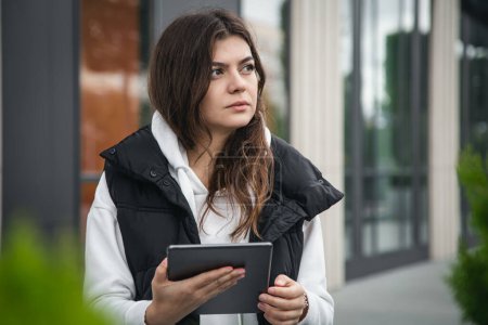 Foto de Mujer joven de negocios se para fuera y utiliza una tableta contra un fondo borroso de un edificio. - Imagen libre de derechos