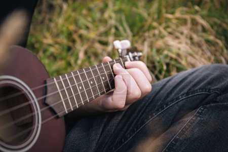 Foto de Un hombre toca la guitarra ukelele en la naturaleza, los dedos de cerca pellizcan las cuerdas, el fondo borroso, el espacio para copiar. - Imagen libre de derechos