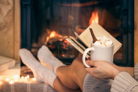 A woman is holding a cup of cocoa with marshmallows and a book while sitting by the fireplace at home.