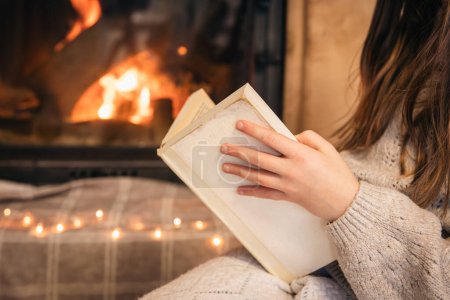 Téléchargez les photos : Gros plan, une petite fille lit un livre sur un fond flou d'une cheminée en feu. - en image libre de droit