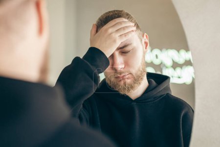 Photo for Sad, tired young man with a beard in front of a mirror, covers his face with his hand, mental health concept. - Royalty Free Image