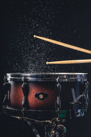 Schlagzeuger mit Schlagstöcken schlägt Snare Drum mit Spritzwasser auf schwarzem Hintergrund.