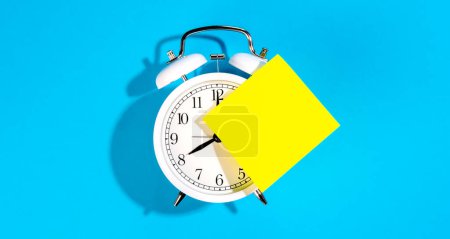 White alarm clock and blank yellow paper sticker on a blue background isolated, flat lay, copy space.