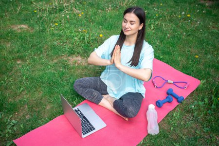 Photo for A young woman meditates after a workout, sitting in front of a laptop on a mat in a green park. - Royalty Free Image