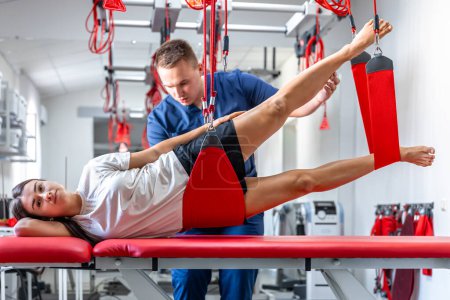 Photo for A young female is doing exercises with special ropes in rehabilitation center, physiotherapist supports his client during workout to recover after the injury. - Royalty Free Image