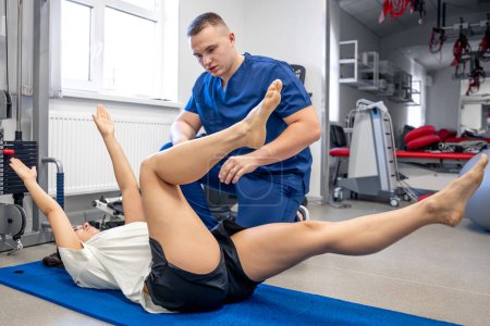 Foto de Mujer acostada en la esterilla de yoga y haciendo gimnasia con la ayuda de su joven fisioterapeuta, concepto de fisioterapia para personas con lesiones. - Imagen libre de derechos