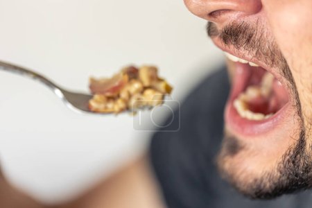 Photo for A hungry man opened his mouth, ready to eat a spoonful of porridge with fruit, close-up. - Royalty Free Image