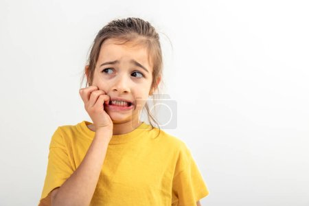 Photo for Portrait of a scared, afraid and anxious little girl in yellow t-shirt on a white background isolated, biting her fingernails, human emotions, copy space. - Royalty Free Image