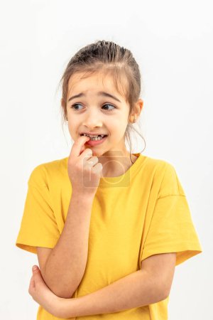 Photo for Portrait of a scared, afraid and anxious little girl in yellow t-shirt on a white background isolated, biting her fingernails, human emotions concept. - Royalty Free Image