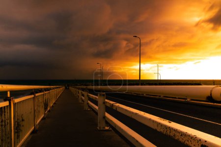 The dark silhouette of a bridge against a fiery sunset over a quiet harbor.