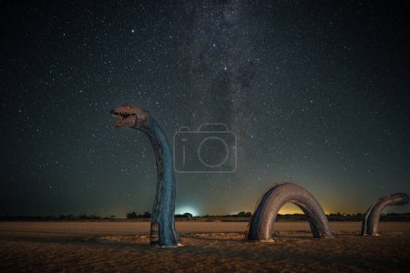An intriguing night landscape featuring a whimsical sculpture of a serpentine creature with its head arching towards the starry sky. The sculpture's textured, scaly surface resembles an otherworldly being merging with the environment. The Milky Way s