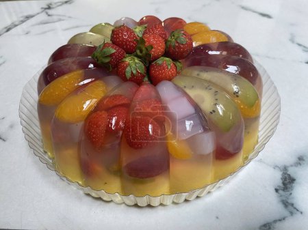 Round shaped healthy fruits jelly pudding on white marble kitchen table. It is made from vanilla jelly, milk, mango juice and filled with fresh strawberry, grape, kiwi, mandarin orange, nata de coco (coconut gel), and fresh strawberry as topping.