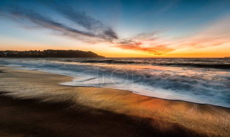Una puesta de sol impresionante proyecta tonos vibrantes a través del océano, como poderosas olas chocan contra la orilla.