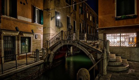 Eine kleine Brücke, die einen schmalen Kanal überspannt und nachts von künstlichen Lichtern beleuchtet wird.