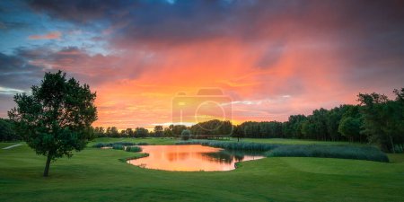 Foto de La foto captura una vista cautivadora de una hermosa puesta de sol que proyecta sus tonos cálidos sobre un campo de golf, destacando un estanque sereno. - Imagen libre de derechos