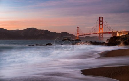 The iconic Golden Gate Bridge seen from the sandy beach, with waves crashing against the shore.